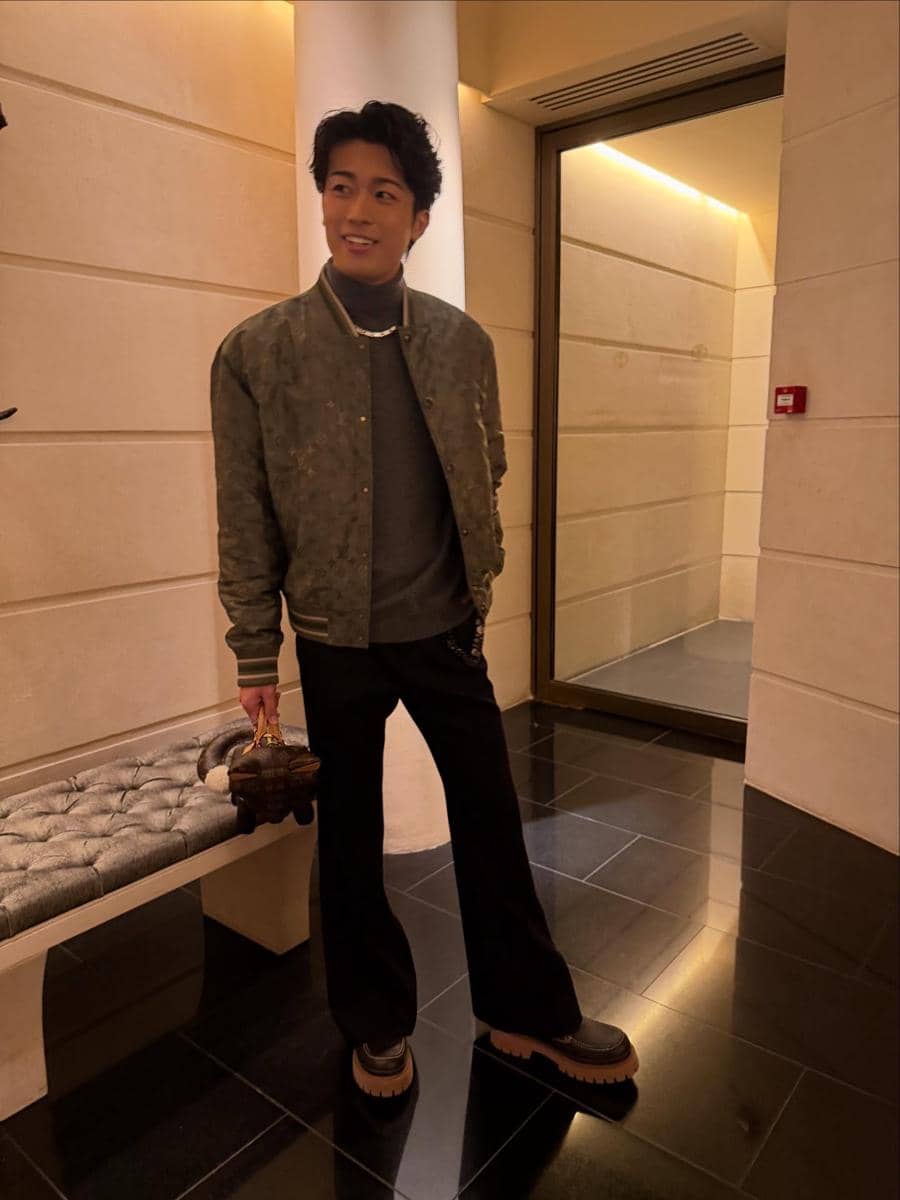 Keito Nakamur berdiri di lorong dengan pencahayaan hangat, mengenakan jaket hijau, celana hitam, dan sepatu tebal sambil memegang tas kecil.