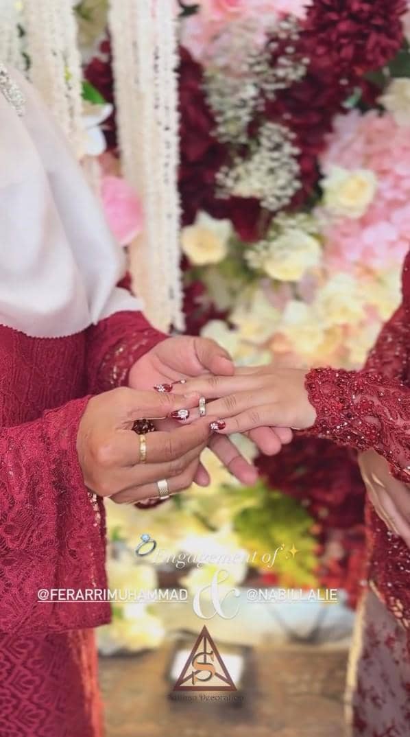 Muhammad Ferarri dan kekasih mengenakan busana merah sedang bertukar cincin lamaran di depan dekorasi bunga berwarna merah muda dan putih.