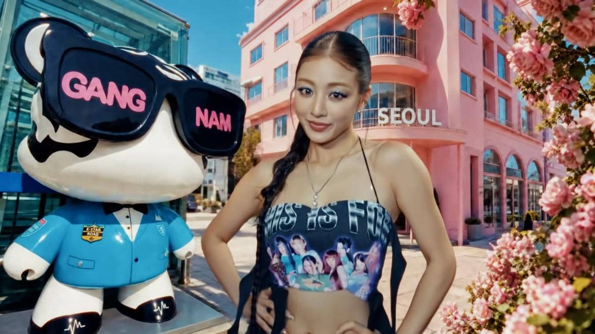 Seorang wanita berpose di depan patung bertuliskan Gangnam dan gedung berwarna merah muda dengan tulisan Seoul di tengah suasana cerah.