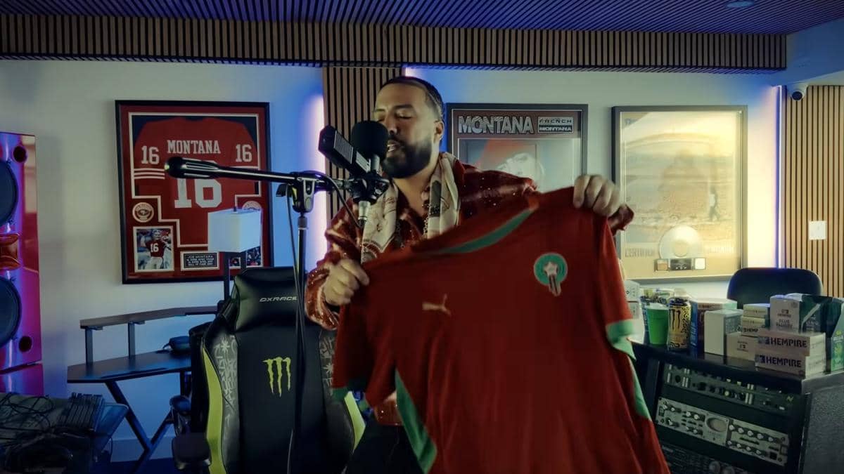 Seorang pria bernyanyi di depan mikrofon di studio rekaman sambil memegang jersey sepak bola berwarna merah dengan logo hijau.