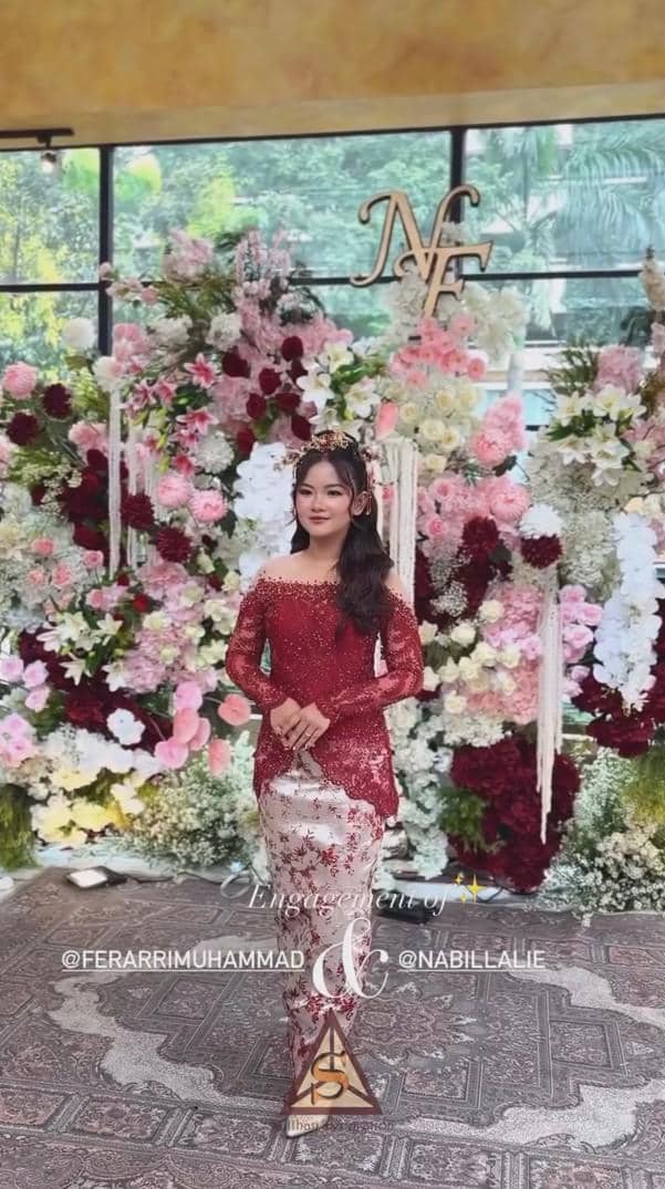 Nabilla mengenakan kebaya merah dan kain batik berdiri di depan dekorasi bunga warna merah muda dan putih dengan inisial NF di atasnya.
