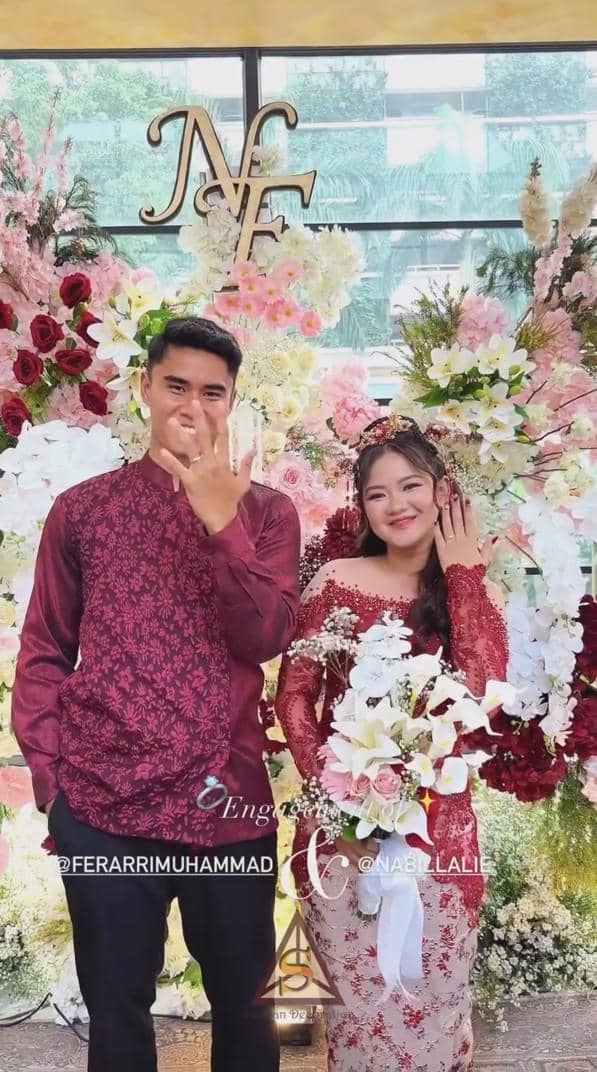 Muhammad Ferarri da kekasih mengenakan busana senada warna merah marun berpose sambil memperlihatkan cincin lamaran di depan dekorasi bunga.