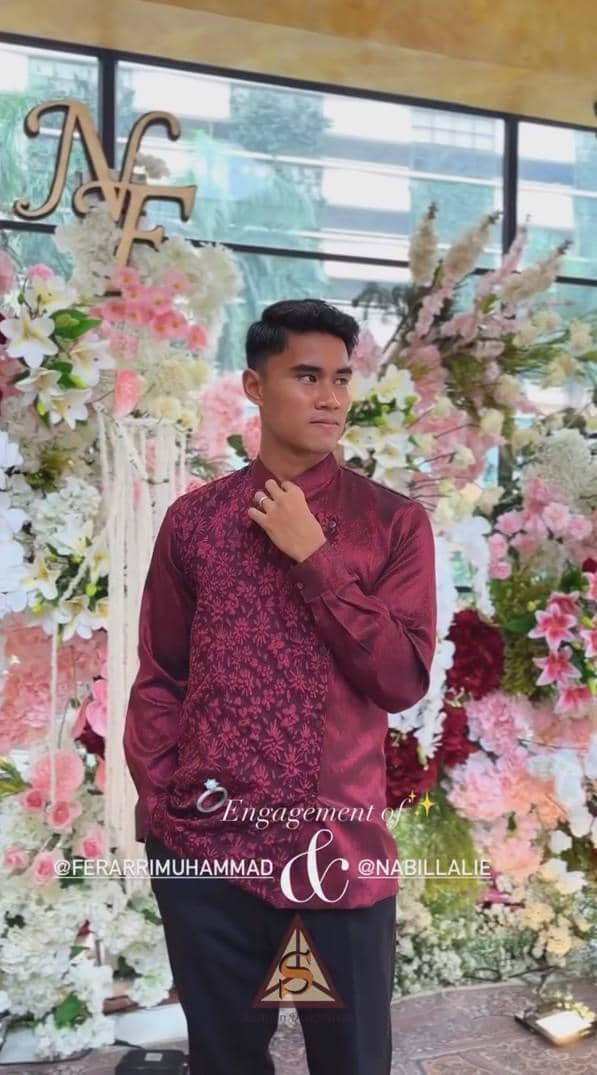 Muhammad Ferarri mengenakan batik merah marun berdiri di depan dekorasi bunga berwarna merah muda dan putih pada acara lamaran.