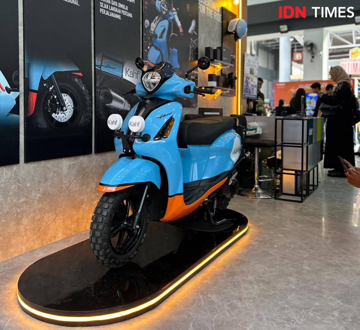 Skuter Yamaha Grand Filano berwarna biru dan oranye hasil modifikasi dipamerkan di atas panggung dengan pencahayaan kuning hangat.