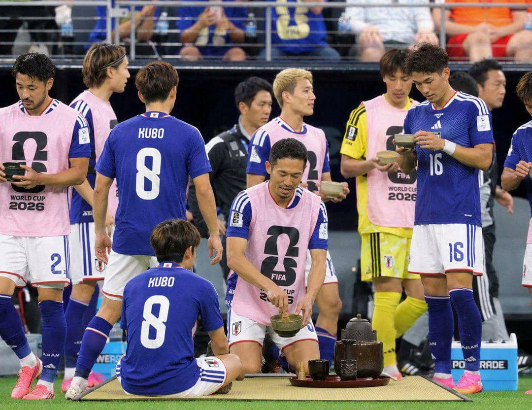 Pemain tim nasional Jepang mengenakan seragam biru dan rompi latihan pink sedang melakukan upacara minum teh di tepi lapangan Piala Dunia 2026.