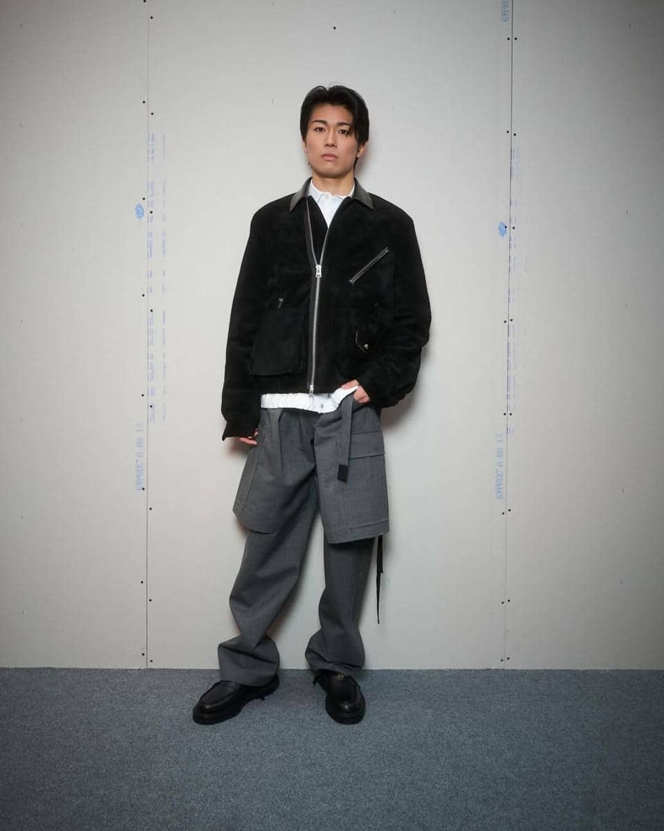 Keito Nakamura berdiri di dalam ruangan dengan dinding polos, mengenakan jaket hitam, kemeja putih, dan celana abu-abu longgar.