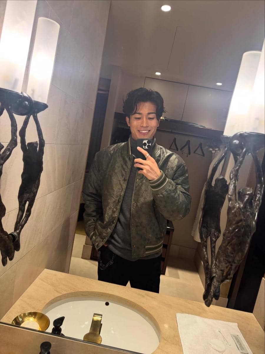 Seorang pria berjaket hijau tersenyum sambil berfoto selfie di depan cermin kamar mandi dengan pencahayaan hangat.