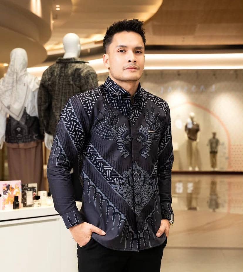 Seorang pria mengenakan batik modern berwarna hitam dengan motif burung di bagian depan, berpose di dalam pusat perbelanjaan.