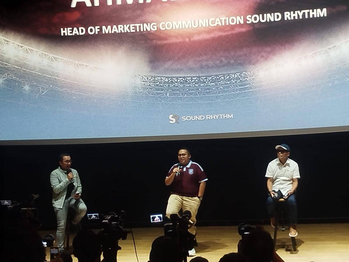 Tiga pria berbicara di atas panggung dalam acara konferensi pers dengan latar layar bertuliskan Sound Rhythm dan stadion di belakangnya.