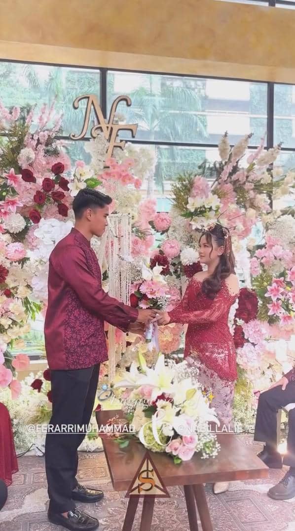 Muhammad Ferarri dan kekasih mengenakan busana merah marun sedang bertunangan di depan dekorasi bunga berwarna merah muda dan putih.