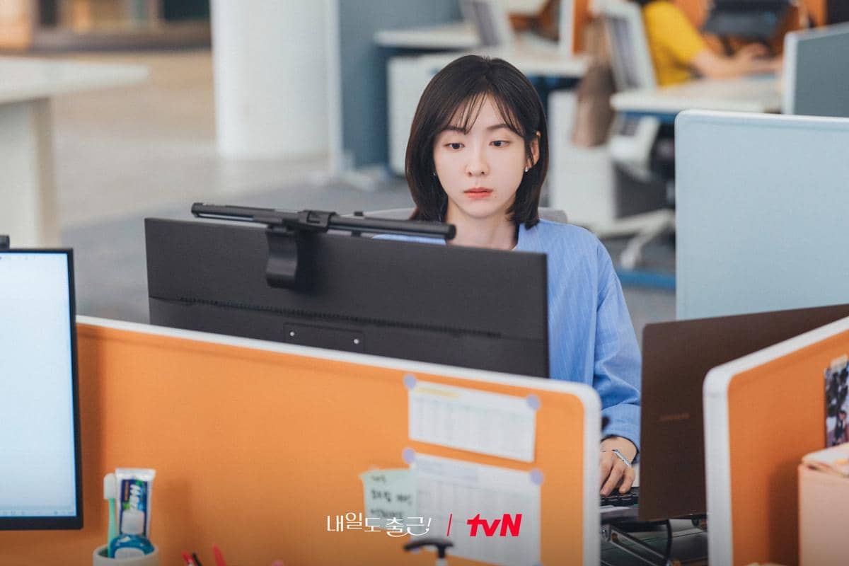 Seorang wanita mengenakan kemeja biru sedang bekerja di depan komputer di kantor terbuka dengan suasana profesional.
