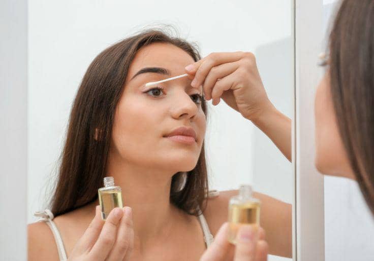 Foto berfokus dekat menampilkan refleksi cermin seorang wanita berambut panjang yang sedang merawat area mata dengan mengoleskan cairan serum dari botol kaca kecil ke bulu matanya menggunakan sebuah korek kuping.