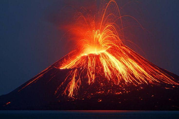 Foto pemandangan malam hari menampilkan siluet sebuah gunung berapi aktif di tengah laut yang sedang meletus kuat meluncurkan pancaran lava pijar merah keemasan dari kawahnya.