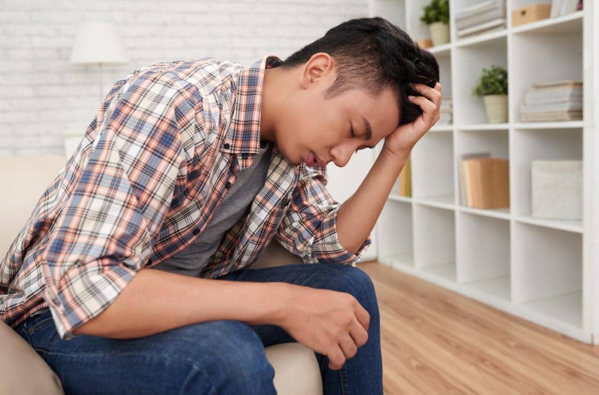 Seorang pria muda duduk di sofa dengan kepala tertunduk dan tangan memegang kepala, tampak sedang menahan perasaan sedih atau stres.