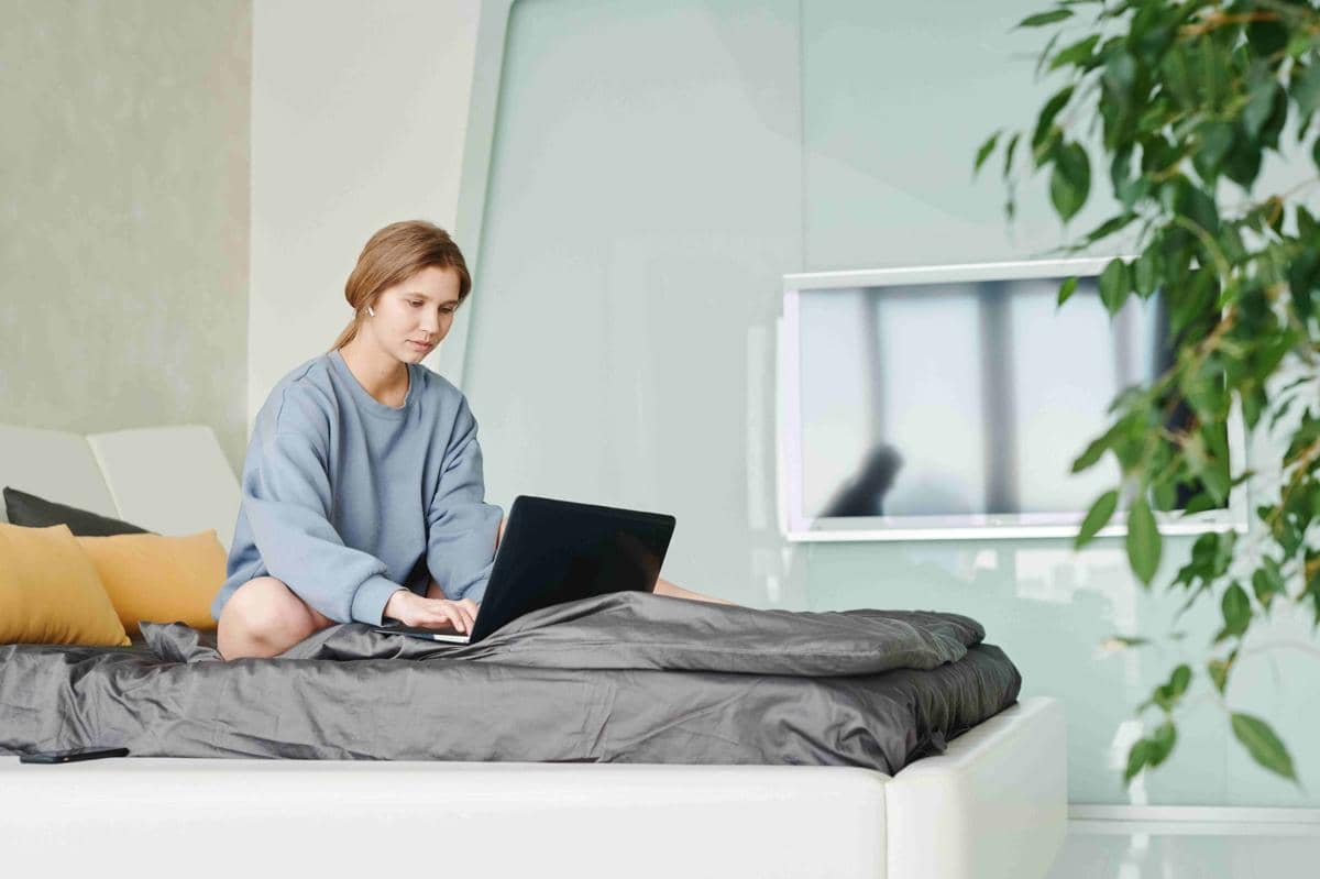 Seorang wanita mengenakan sweater abu-abu bekerja menggunakan laptop di atas tempat tidur dengan bantal berwarna kuning dan abu-abu.
