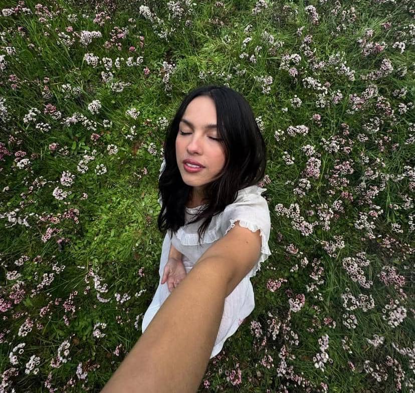 Seorang perempuan berambut hitam mengenakan gaun putih berpose dengan mata terpejam di tengah padang rumput berbunga kecil berwarna merah muda.