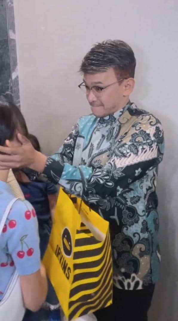 Ruben onsu mengenakan batik bermotif biru memegang wajah Thalia sambil membawa tas belanja kuning bergaris hitam.