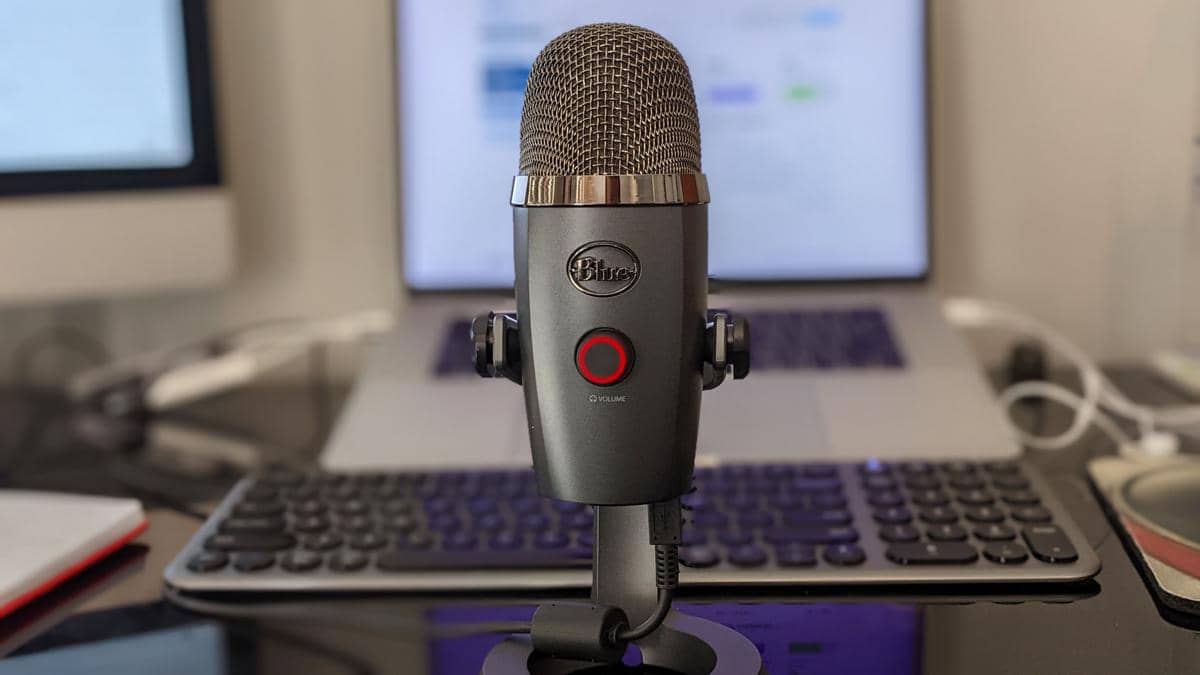Mikrofon podcast berwarna abu-abu berdiri di atas meja kerja dengan laptop dan keyboard di latar belakang.