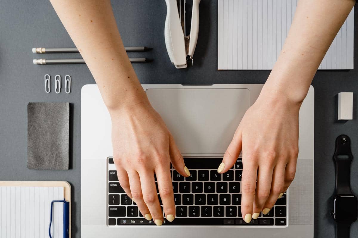 Tangan seseorang sedang mengetik di laptop dengan perlengkapan kerja seperti pensil, kertas, dan jam tangan di meja.