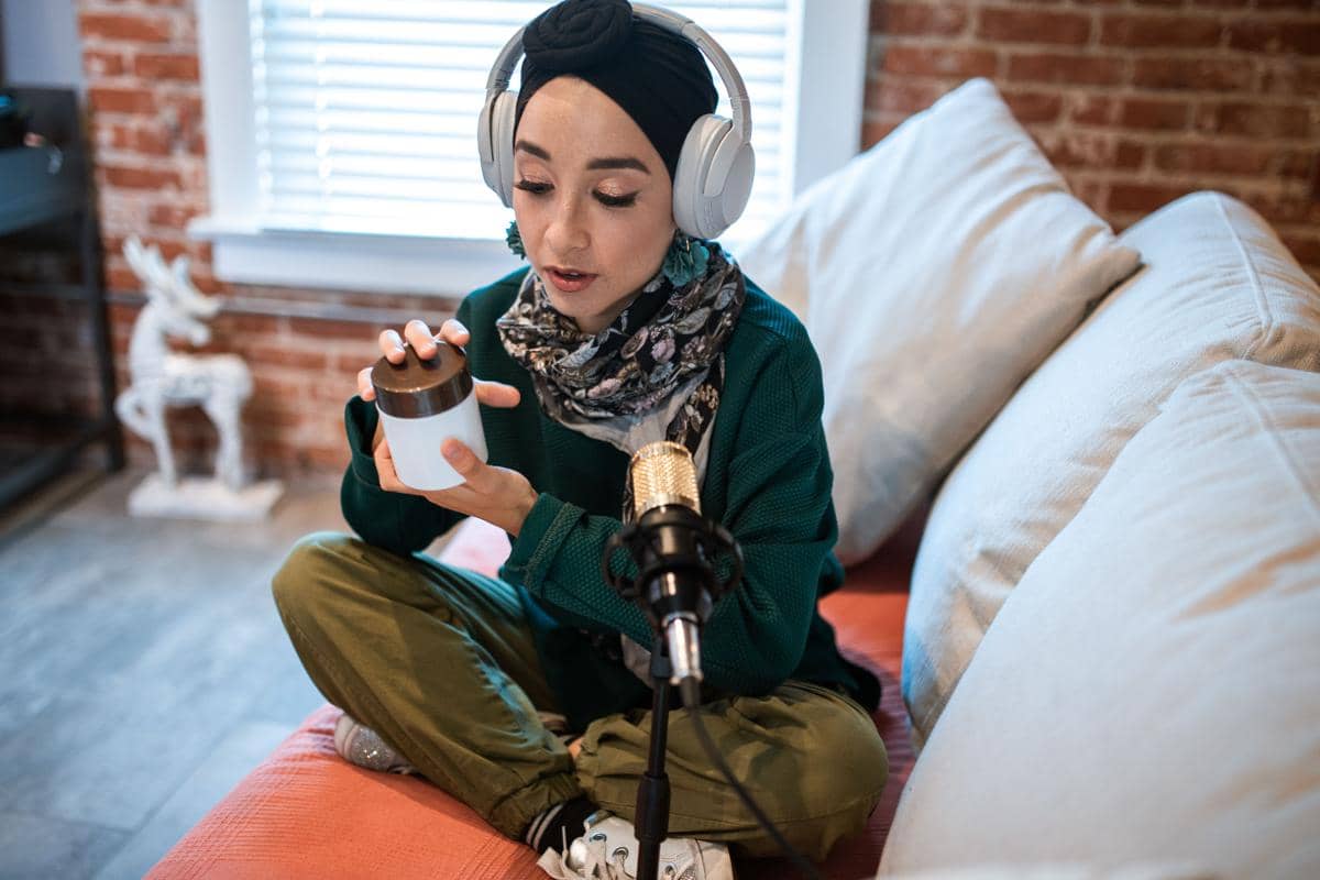 Seorang perempuan berhijab mengenakan headphone sedang berbicara di depan mikrofon sambil memegang wadah kecil di ruang santai.