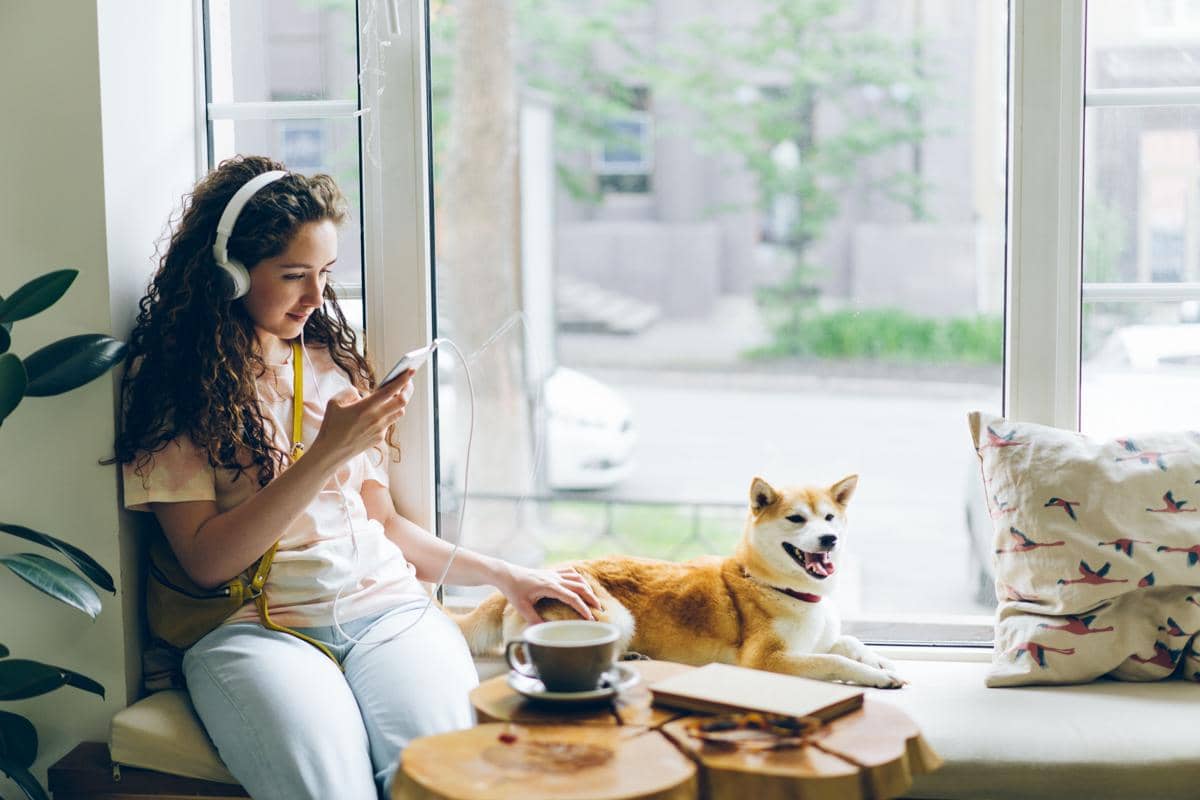 Seorang wanita duduk di dekat jendela sambil mendengarkan musik dengan headphone dan menemani seekor anjing yang berbaring santai.