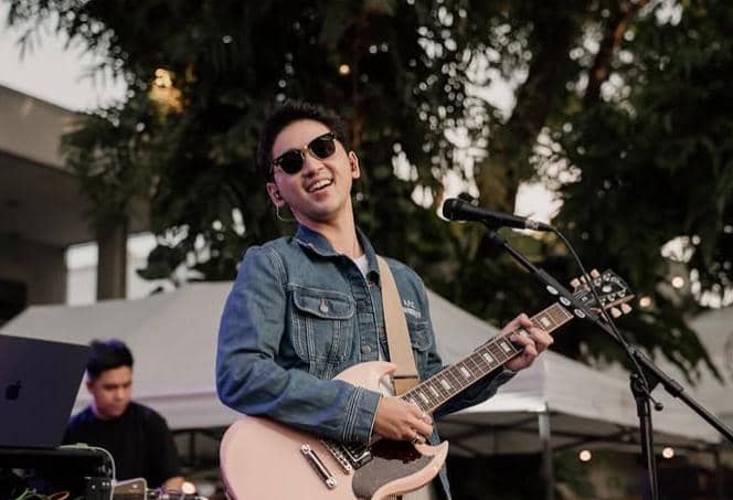 Seorang pria mengenakan jaket denim dan kacamata hitam sedang memainkan gitar listrik di atas panggung luar ruangan.