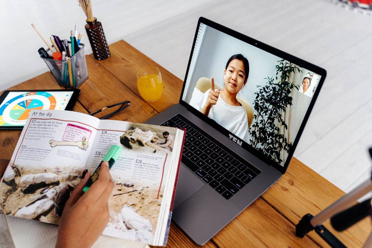 Seseorang mengikuti kursus online melalui laptop sambil mencatat di buku dan menyorot teks dengan stabilo di meja kayu.