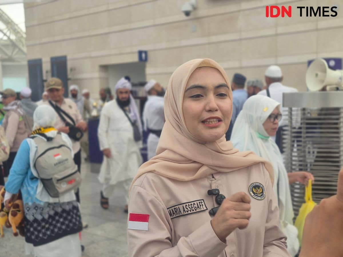Seorang perempuan berjilbab krem mengenakan seragam Kemenhaj berbicara di area keberangkatan haji yang ramai dengan jemaah.
