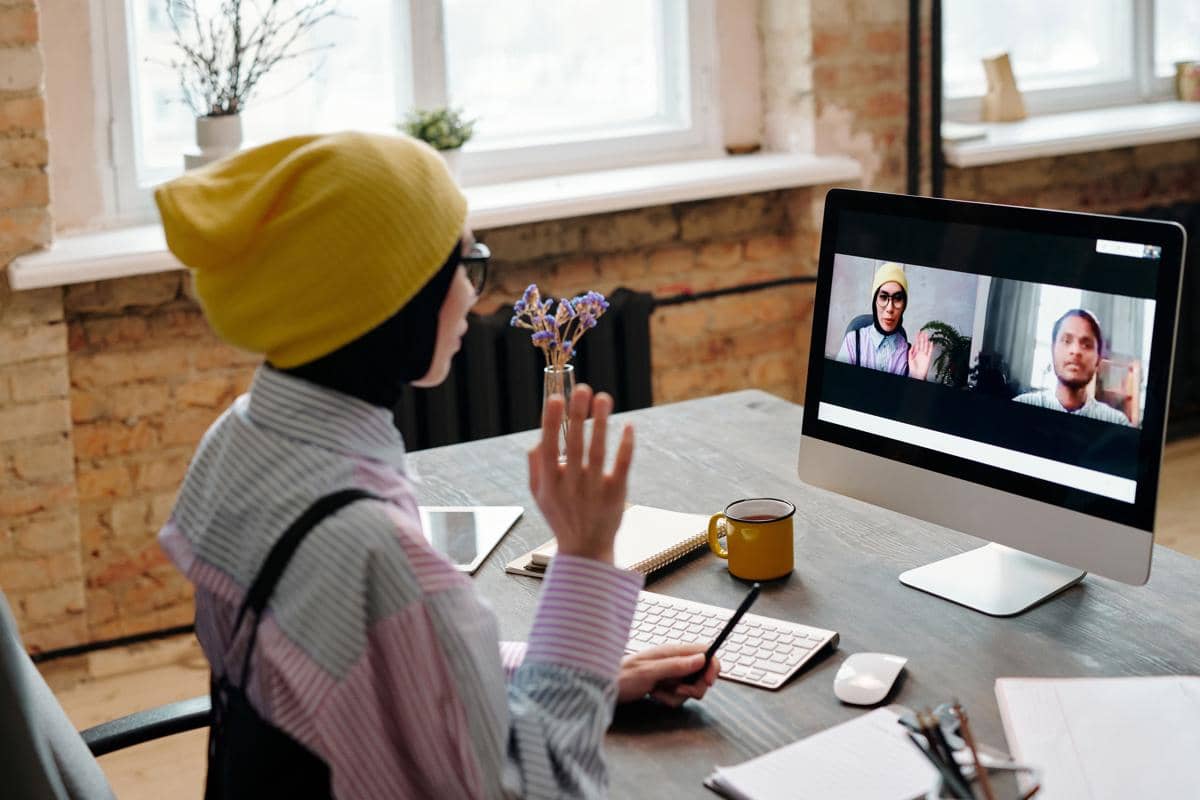 Seorang perempuan mengenakan beanie kuning sedang melakukan panggilan video dengan dua orang di layar komputer di meja kerja.