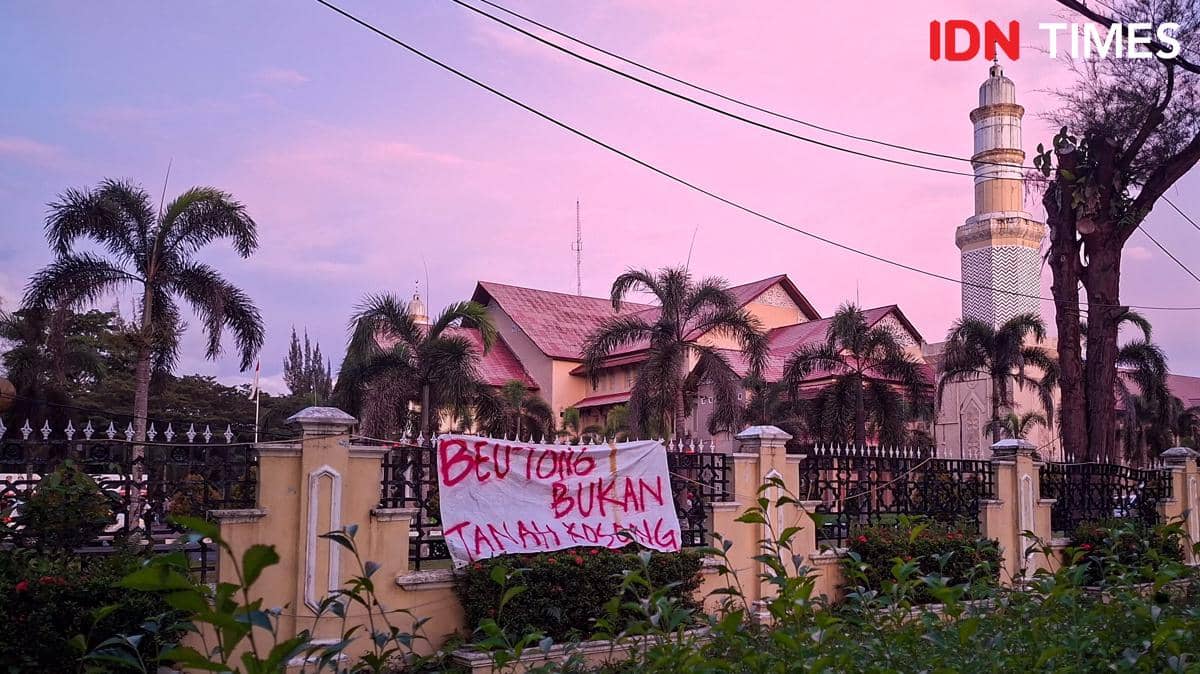 Spanduk bertuliskan pesan protes terpasang di pagar depan bangunan bercat kuning dengan latar langit senja di Beutong Ateuh Banggalang.