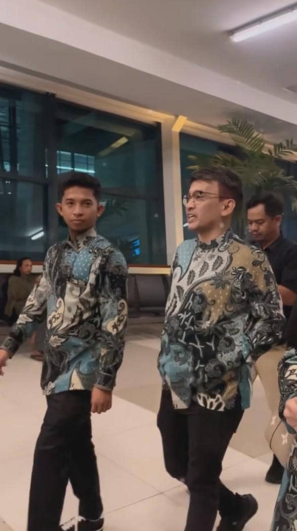 Ruben Onsu mengenakan batik berwarna biru dan hitam berjalan bersama rombongan di area bandara dengan suasana malam.