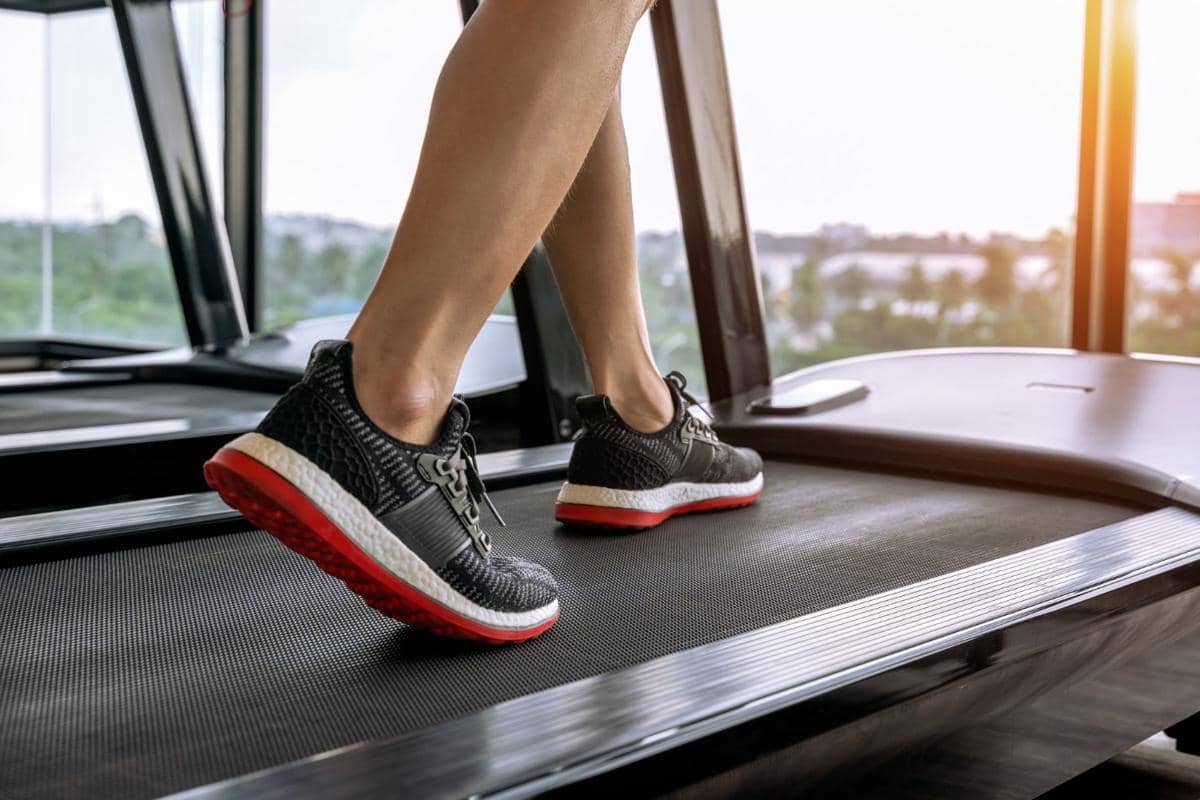 Seseorang mengenakan sepatu olahraga hitam dengan sol merah sedang berjalan di atas walking pad di dalam ruangan dengan cahaya matahari masuk.