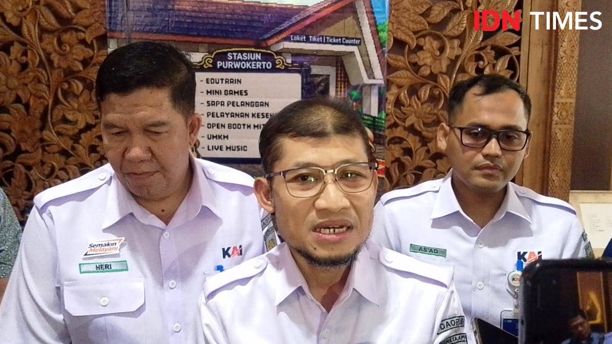 Tiga petugas KAI mengenakan seragam putih sedang memberikan keterangan di Stasiun Purwokerto dengan latar ornamen kayu dan papan informasi.