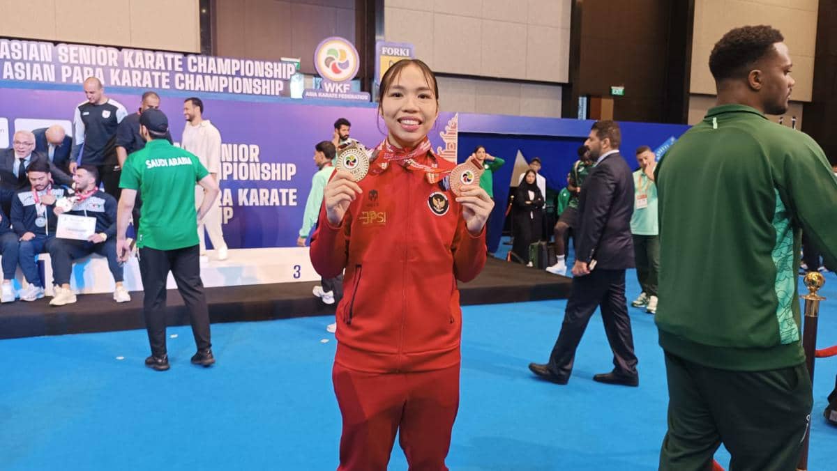 Atlet karate perempuan Indonesia mengenakan jaket merah memegang dua medali di arena Kejuaraan Karate Asia dengan latar peserta lain.