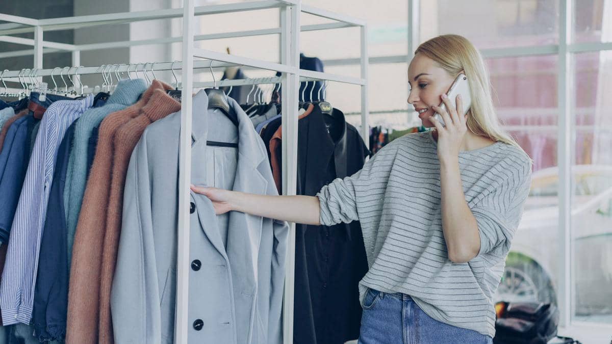Seorang wanita berbicara di telepon sambil memilih pakaian di rak baju, menggambarkan aktivitas bisnis preloved di butik modern.