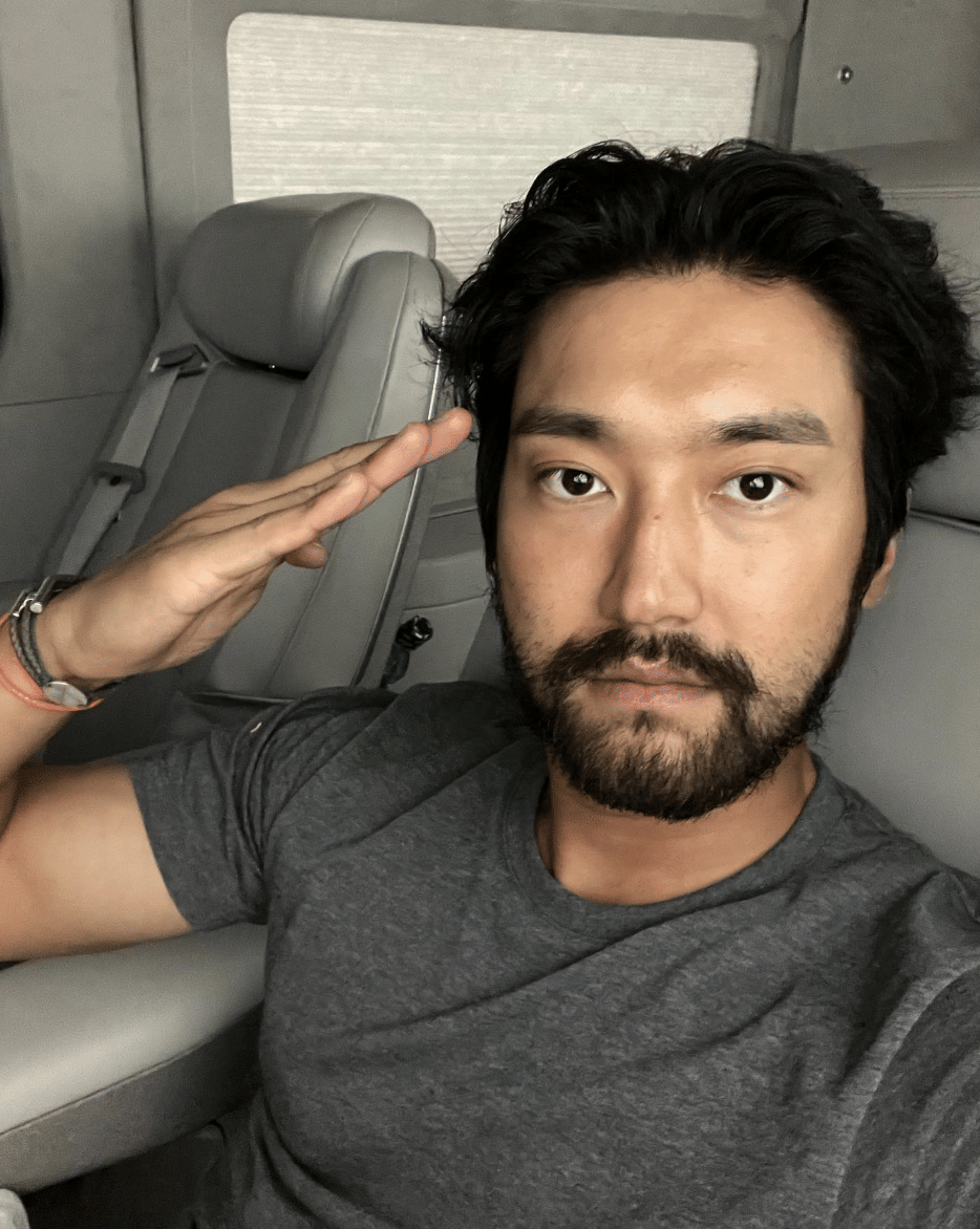 Seorang pria berjanggut mengenakan kaus abu-abu duduk di kursi mobil sambil memberi salam dengan tangan kanan di dekat kepala.
