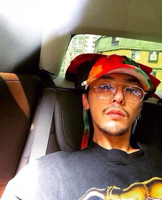 Seorang pria mengenakan kacamata bulat dan topi berwarna merah hijau duduk di kursi belakang mobil dengan cahaya matahari masuk dari jendela.