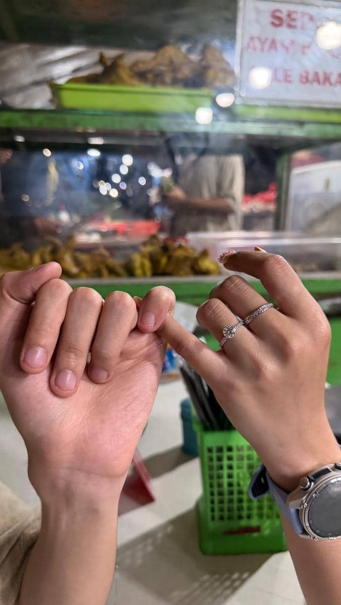 Dua tangan saling berjanji kelingking, salah satunya memakai cincin berlian di depan gerobak makanan malam hari.