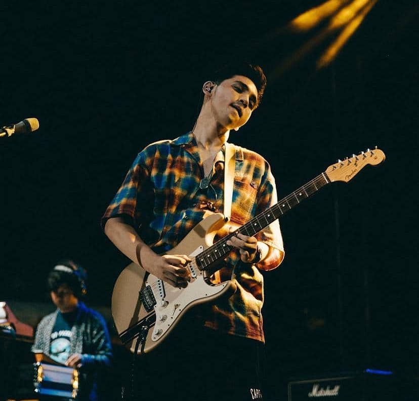 Seorang pria mengenakan kemeja bermotif warna-warni sedang memainkan gitar listrik di atas panggung dengan pencahayaan panggung hangat.