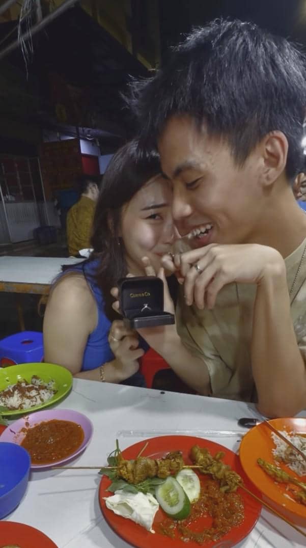 Yuka dan Michelle tersenyum bahagia di warung makan malam hari, sambil memperlihatkan kotak cincin pertunangan di meja penuh hidangan.