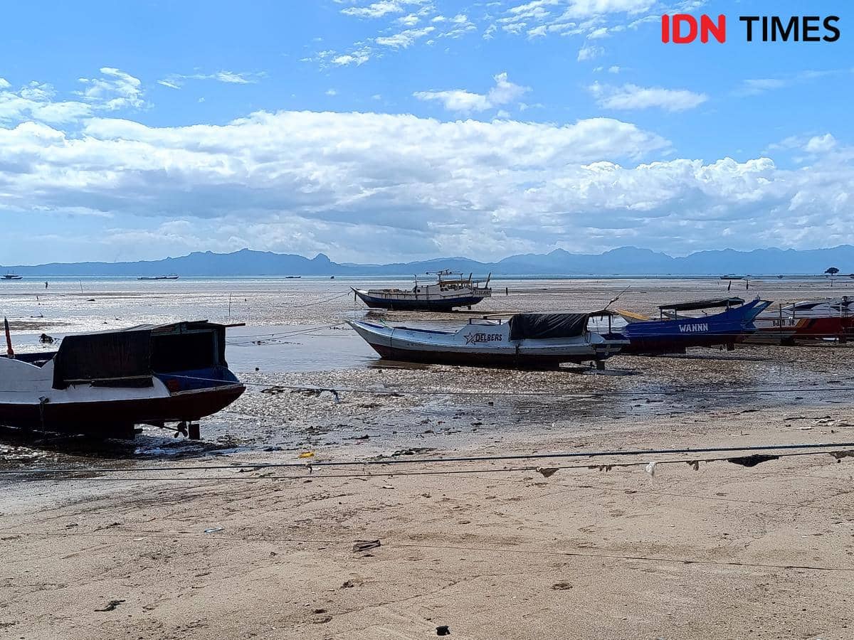 Beberapa kapal nelayan bersandar di tepi Pantai Oesapa saat air laut surut dengan latar langit biru dan pegunungan di kejauhan.