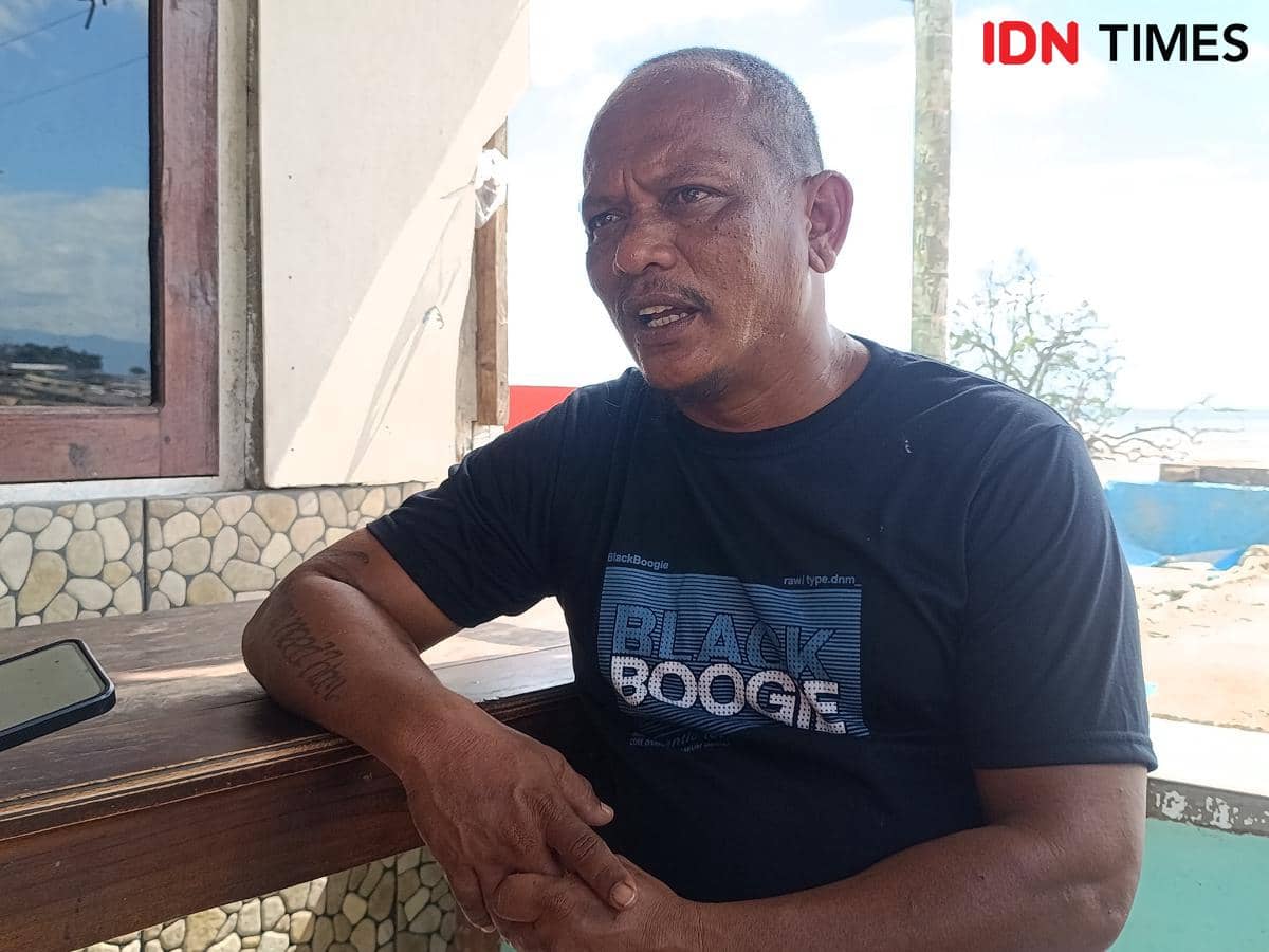 Seorang pria duduk di meja kayu mengenakan kaus hitam bertuliskan 'Black Boogie', berbicara dengan latar belakang area terbuka dekat pantai.