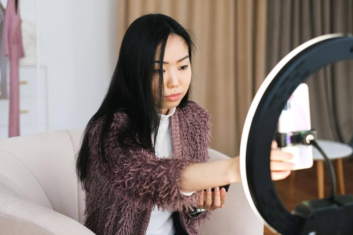 Seorang perempuan sedang menyiapkan ponsel di depan ring light untuk membuat konten video di dalam ruangan.