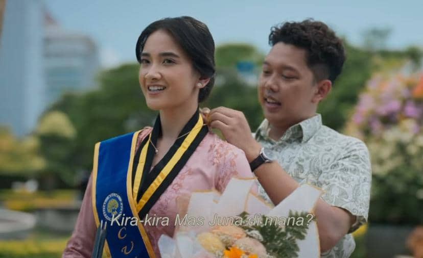 Seorang pria memasangkan medali pada wanita berbusana kebaya dengan selempang wisuda di luar ruangan yang dipenuhi bunga.