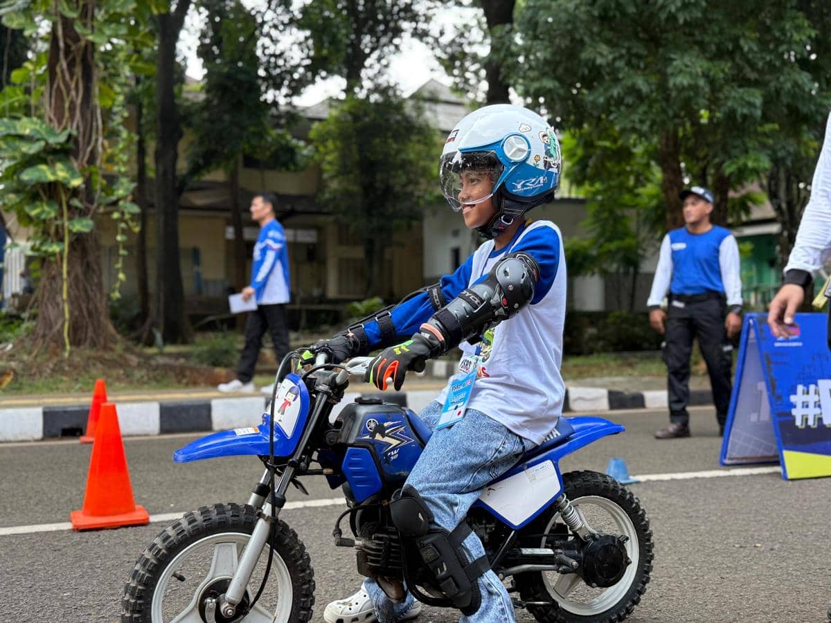 Seorang anak mengenakan helm dan pelindung tubuh saat belajar mengendarai motor mini biru di area latihan safety riding.