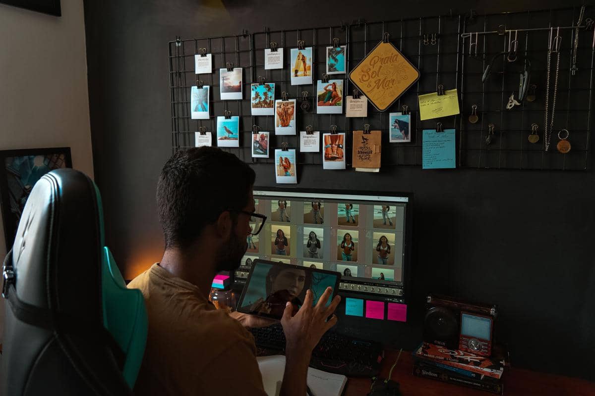 Seorang pria sedang mengedit foto di komputer dengan tablet di tangannya, di ruang kerja dengan dinding berisi foto-foto dan catatan.