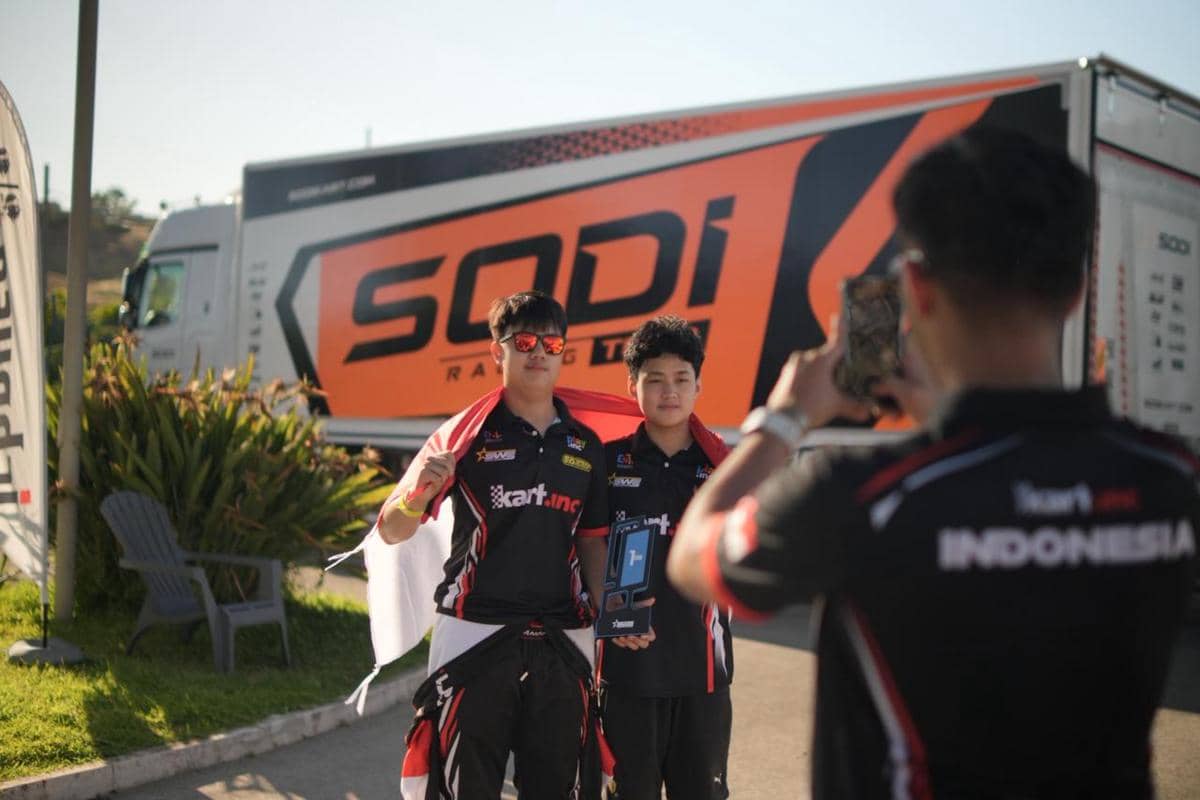 Dua pembalap muda mengenakan seragam tim karting Indonesia berpose sambil memegang trofi di depan truk bertuliskan Sodi Racing Team.