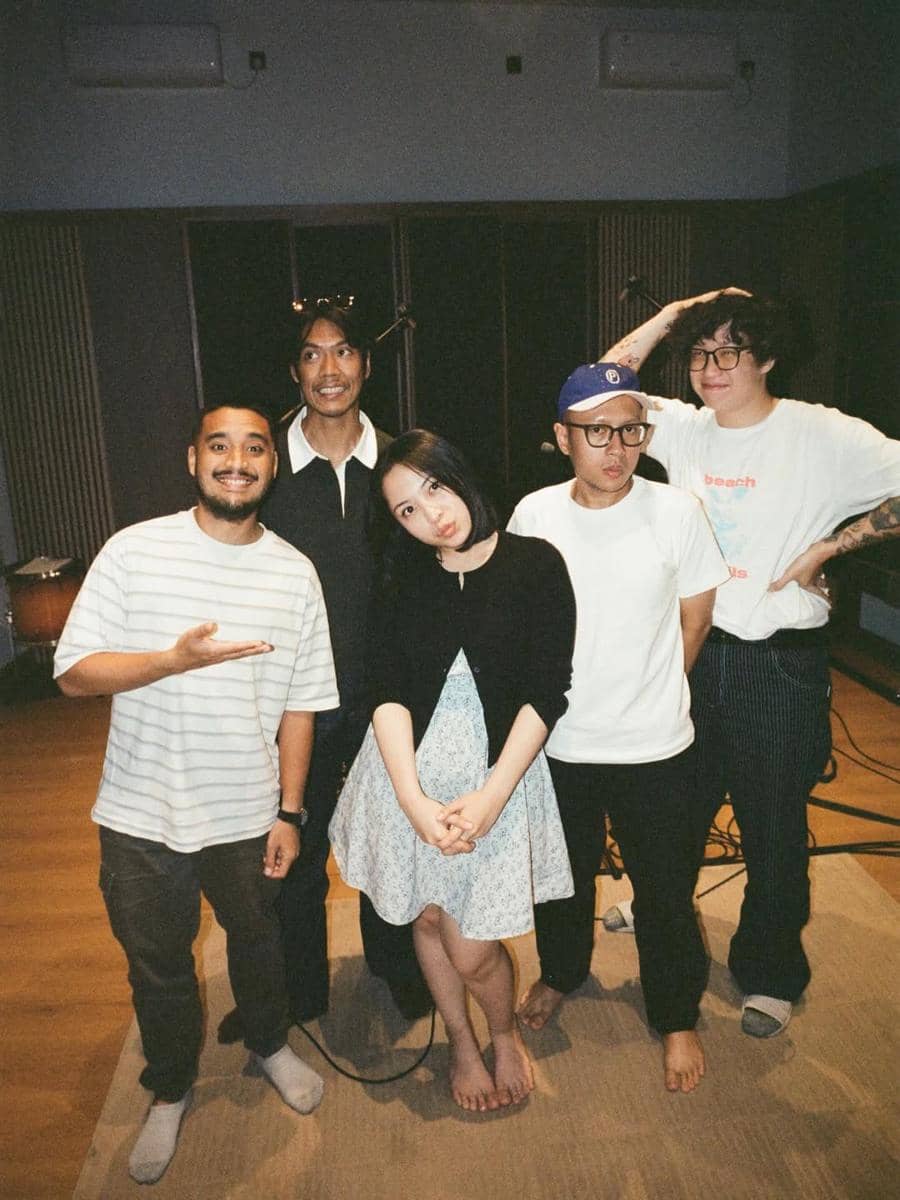 Lima orang berdiri bersama di dalam studio musik dengan suasana santai, mengenakan pakaian kasual dan tersenyum ke arah kamera.