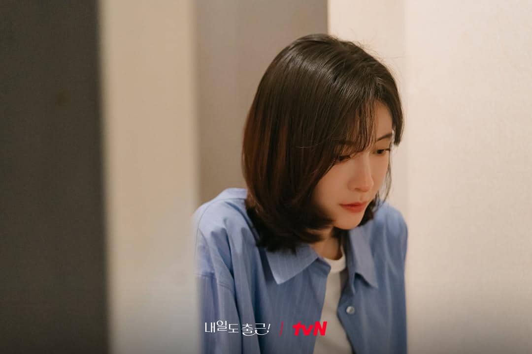 Seorang wanita berambut pendek mengenakan kemeja biru tampak duduk dengan ekspresi serius dalam cuplikan drama Korea See You at Work Tomorrow.