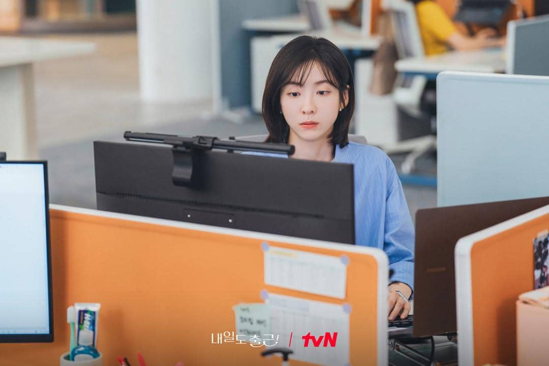 Seorang wanita mengenakan kemeja biru sedang fokus bekerja di depan komputer di ruang kantor modern dengan suasana profesional.